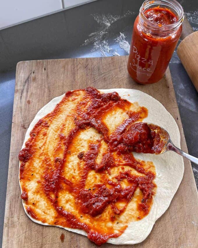 A spoon spreads tomato pizza sauce from a jar onto a round, uncooked pizza dough on a wooden board.