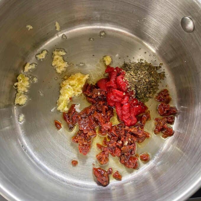 Chopped sun-dried tomatoes, tomato paste, minced garlic, and dried herbs in a stainless steel pot.