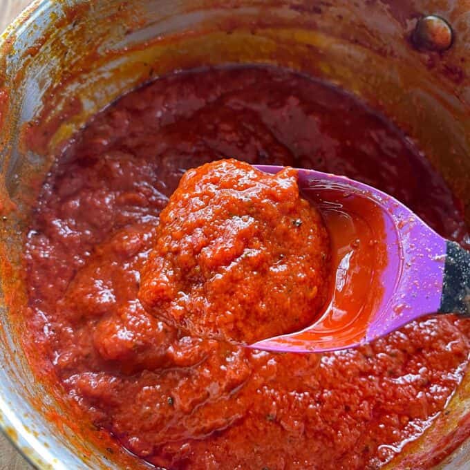 A close-up of thick tomato sauce being stirred with a purple spoon in a metal pot.