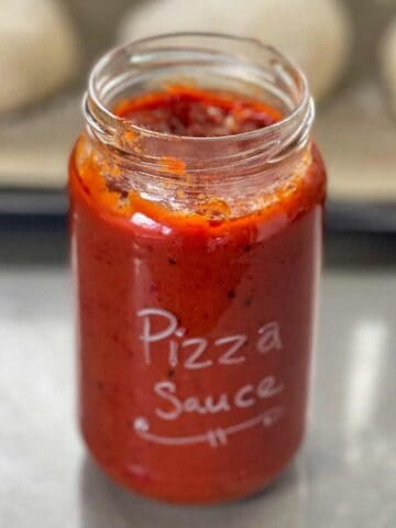 A glass jar labeled Pizza Sauce filled with red tomato sauce sits on a metal surface, with blurred dough balls in the background.