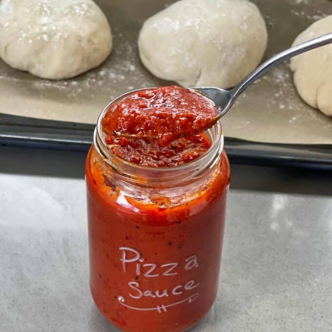 A spoonful of red pizza sauce is held over an open jar labeled Pizza Sauce, with dough balls on a baking tray in the background.