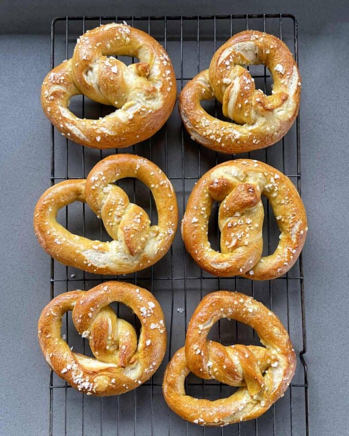 Six salted soft pretzels cooling on a metal rack placed on a gray surface.