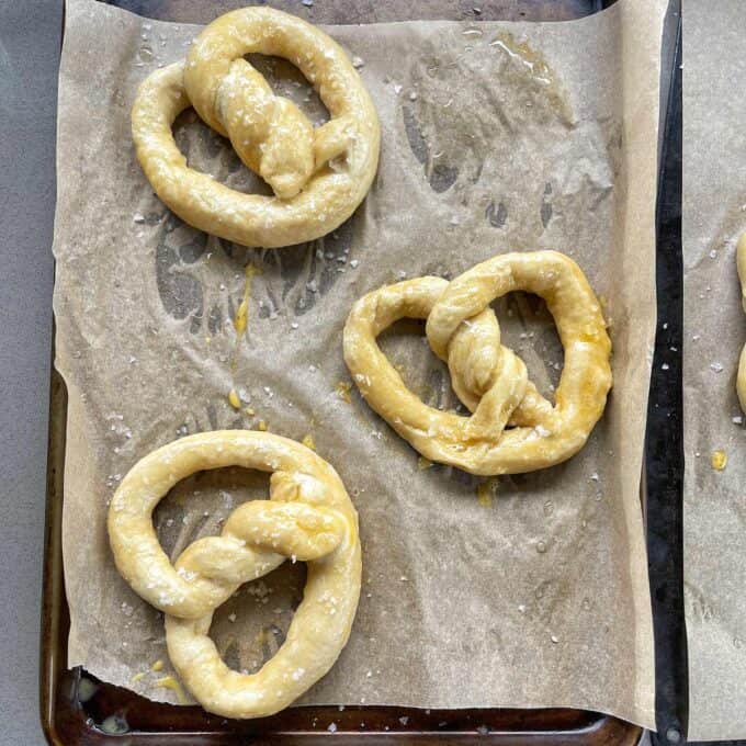 Three unbaked pretzels sprinkled with coarse salt are arranged on a parchment-lined baking sheet.