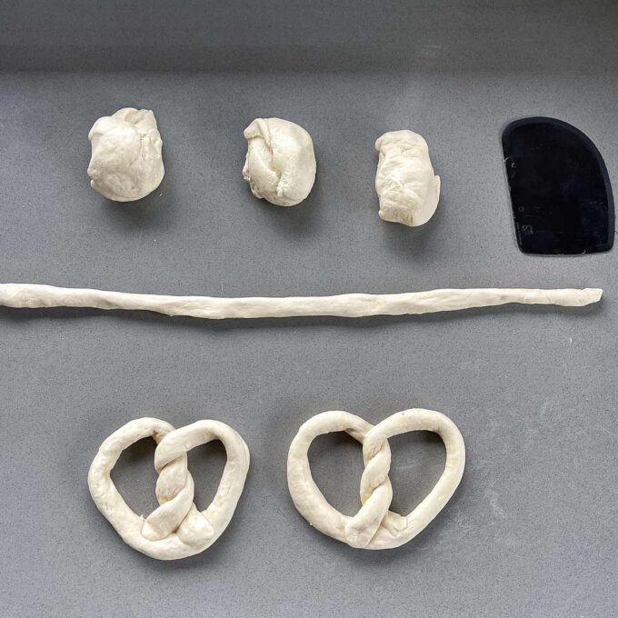 Five pieces of dough in various stages: balls, a rolled rope, and two shapes formed into classic pretzel loops, on a gray surface with a black dough scraper.