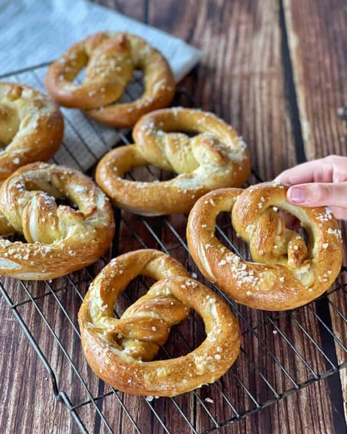 Several salted soft pretzels rest on a wire cooling rack, with a hand picking up one pretzel. The rack is placed on a wooden surface.