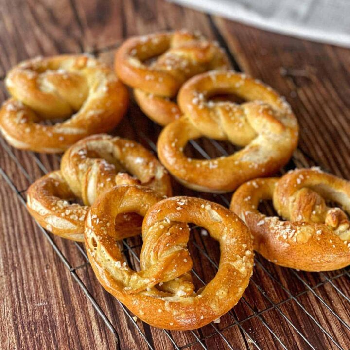 Seven golden-brown soft pretzels sprinkled with coarse salt are cooling on a wire rack atop a wooden surface.