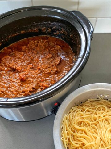 A slow cooker filled with meat sauce sits on a counter next to a colander containing cooked spaghetti noodles.