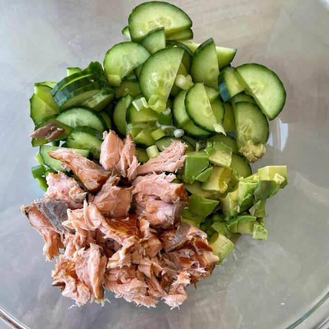 A glass bowl with sliced cucumber, diced avocado, chopped green onion, and flaked cooked salmon arranged in separate sections.