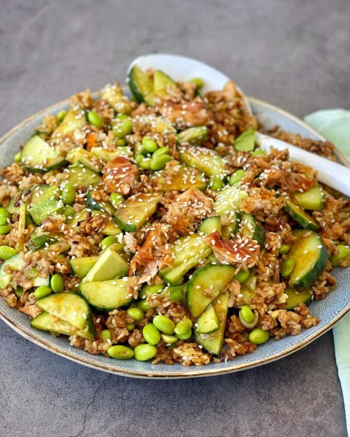 A plate of fried rice mixed with sliced cucumber, edamame, shredded fish, and sprinkled with sesame seeds, served with a white spoon on the side.