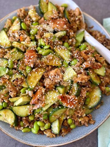 A plate of salad featuring rice, cucumbers, edamame, avocado, and shredded protein topped with sesame seeds, served with chopsticks on the side.
