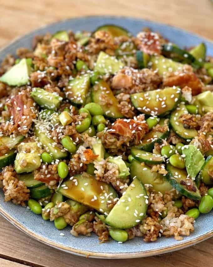 A plate of rice salad topped with cucumbers, edamame, avocado, pieces of fish, and sprinkled with sesame seeds.