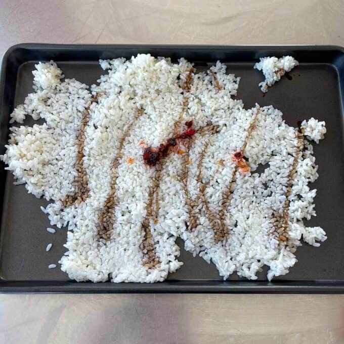 A baking tray with a spread-out layer of cooked white rice, streaked with lines of soy sauce and a small amount of chili sauce.