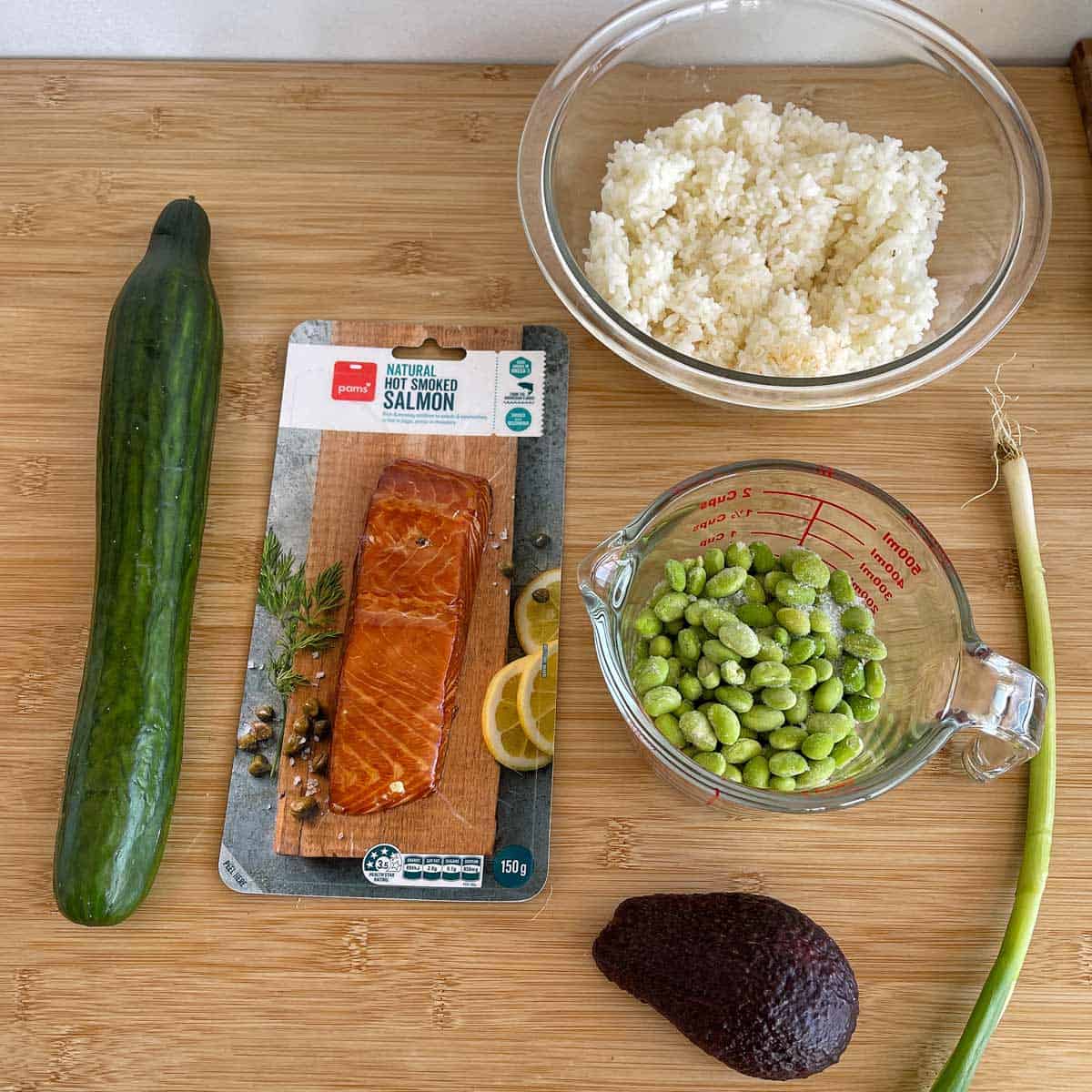 A cucumber, packaged smoked salmon, bowl of rice, measuring cup of edamame, avocado, and green onion arranged on a wooden surface.