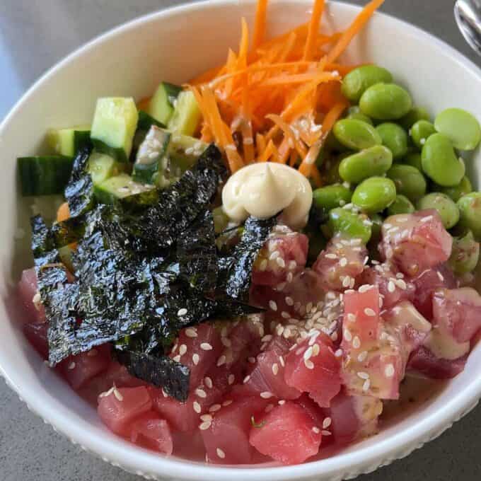 A poke bowl containing diced raw tuna, seaweed, edamame, shredded carrots, cucumber, a dollop of mayo, and sesame seeds, arranged in sections in a white bowl.