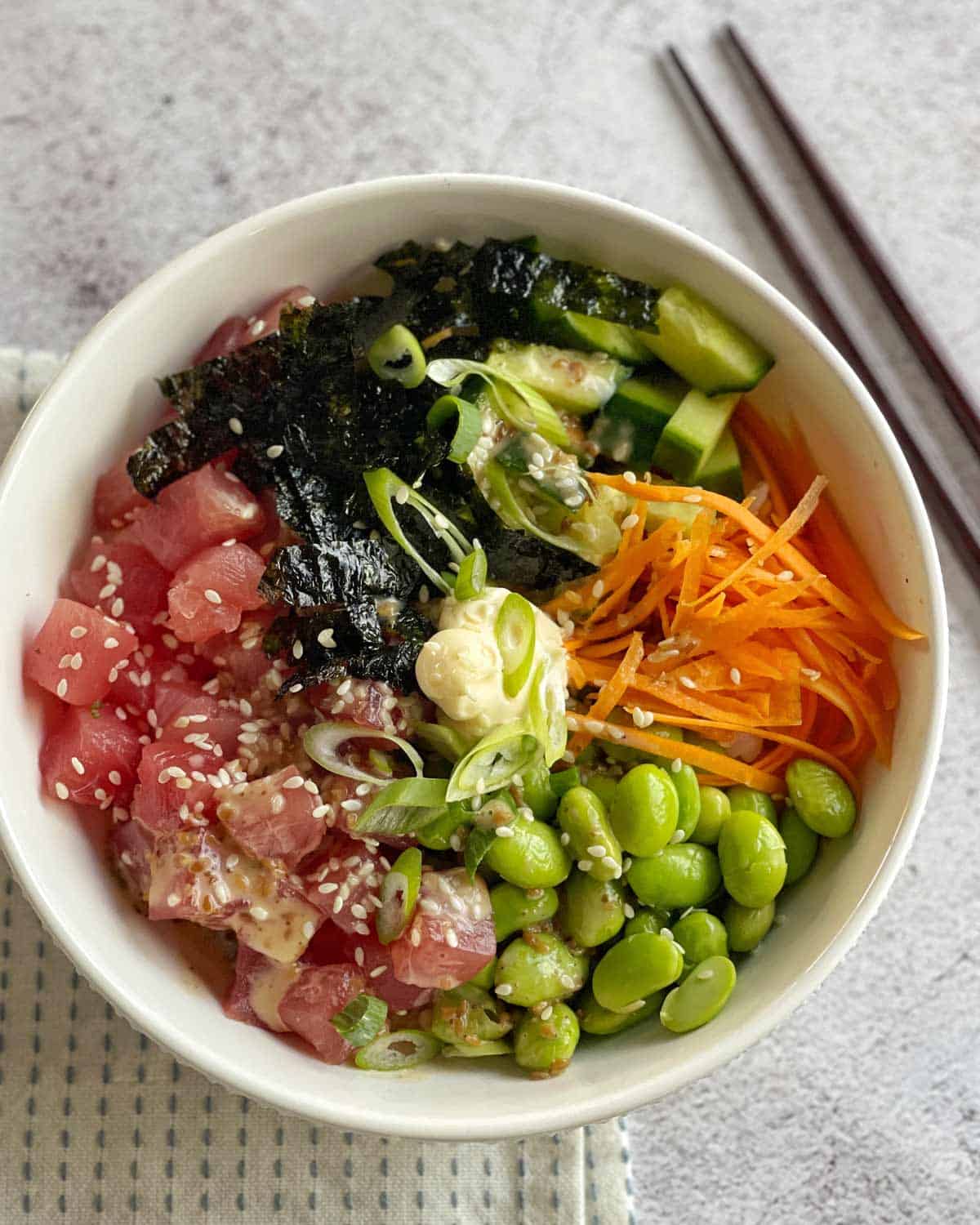 Fresh Tuna Poke Bowls A bowl of poke with diced raw fish, seaweed, cucumber, shredded carrots, edamame, sesame seeds, green onions, and a dollop of sauce, next to a pair of chopsticks.