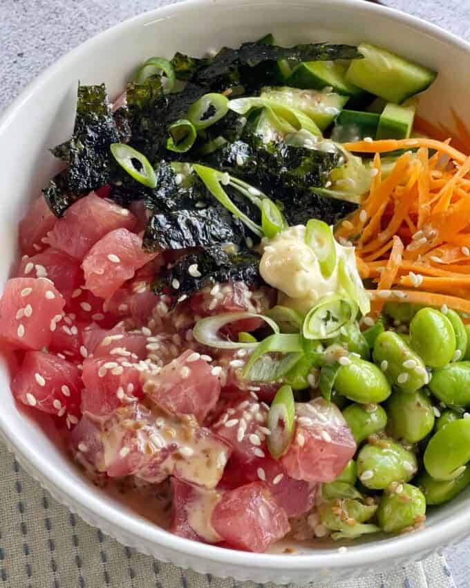 A poke bowl with diced raw fish, edamame, shredded carrots, cucumber, seaweed, green onions, sesame seeds, and a drizzle of sauce in a white bowl.