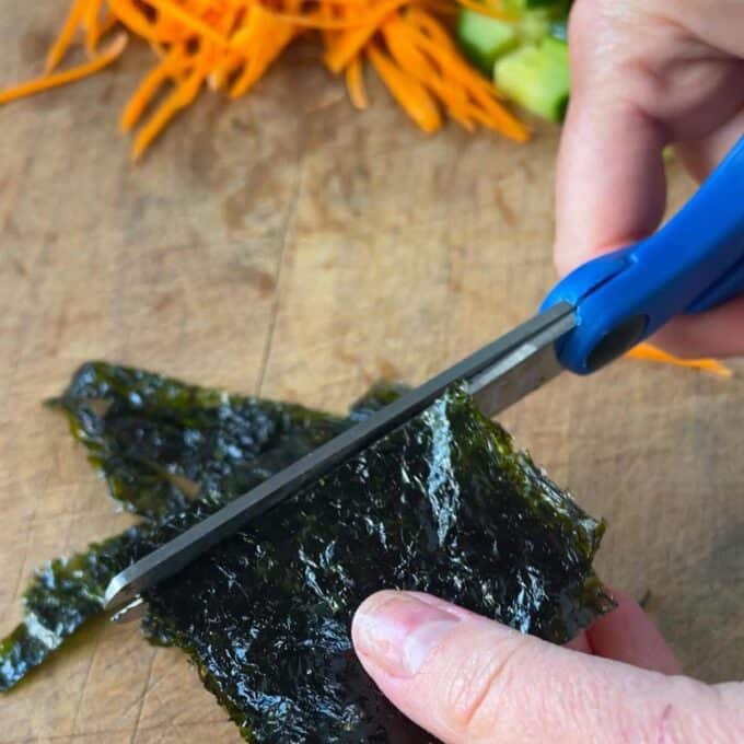 A hand uses blue-handled scissors to cut a sheet of seaweed on a wooden board, with sliced vegetables in the background.