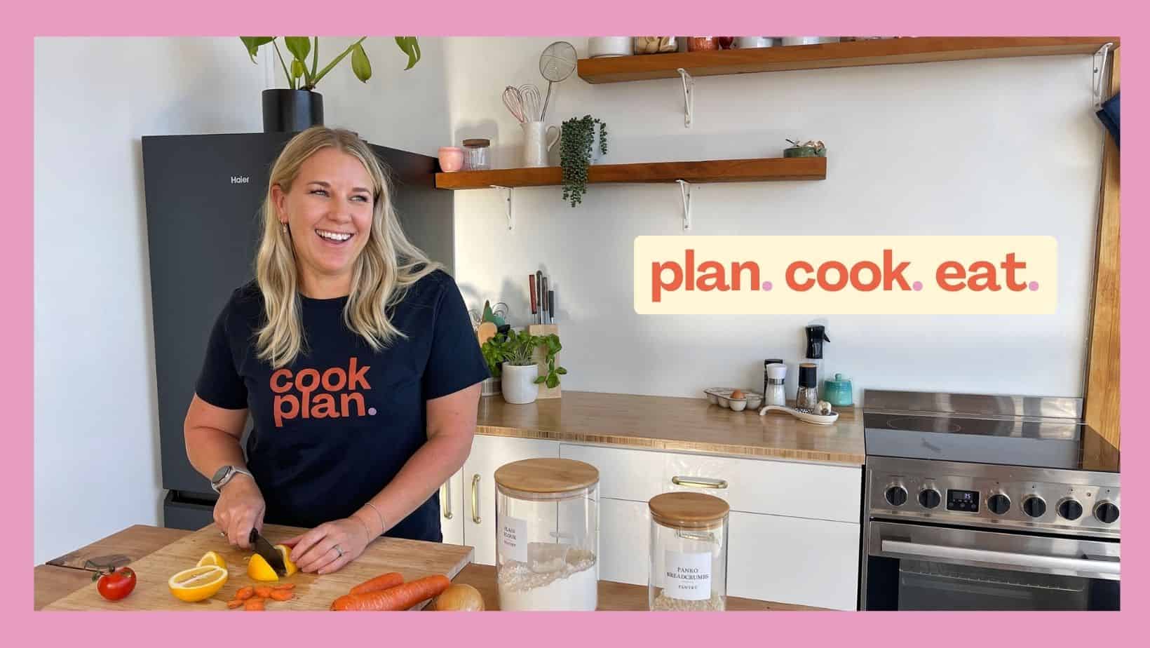 A woman smiles while chopping vegetables in a modern kitchen. She wears a shirt that says cook. plan. with a sign in the background reading plan. cook. eat.