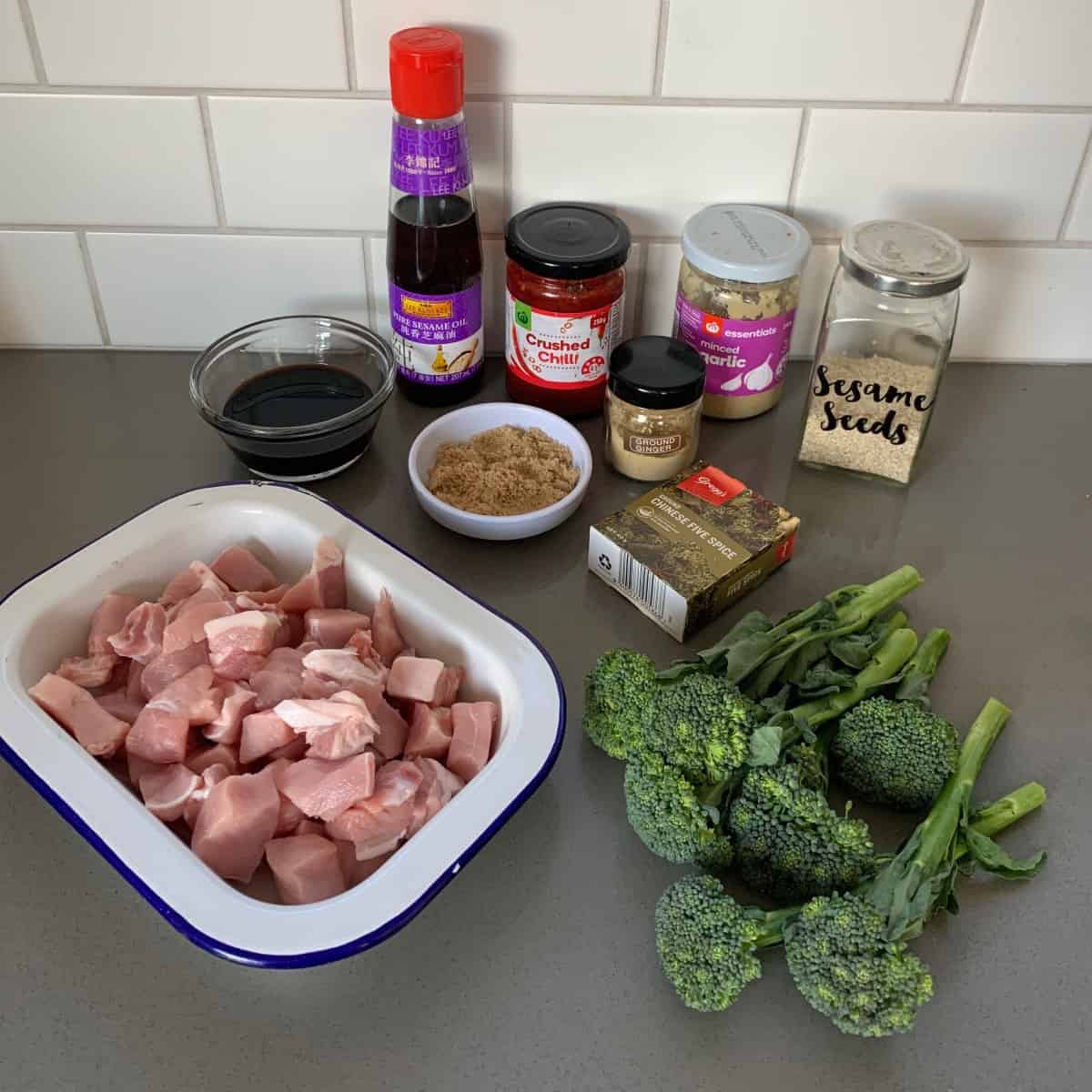 Chopped raw pork, broccoli, and various seasonings and sauces, including soy sauce, brown sugar, sesame seeds, and chili sauce, are arranged on a kitchen counter.