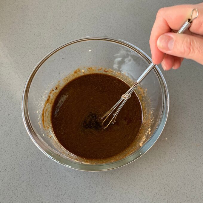 A hand uses a whisk to mix a dark brown liquid in a clear glass bowl on a gray countertop.