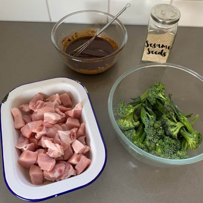 Ingredients for a meal: raw diced chicken in a tray, cooked broccoli in a bowl, a bowl of brown sauce with a whisk, and a jar labeled Sesame Seeds on a kitchen counter.