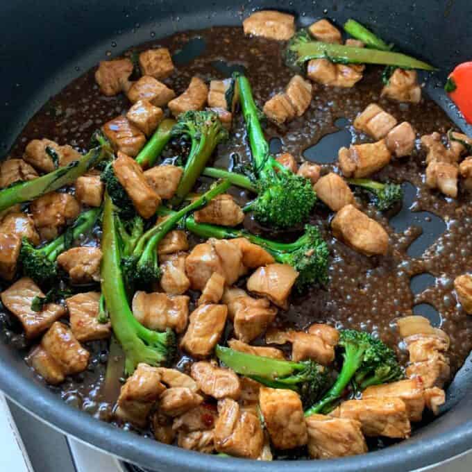 Stir-fry with pieces of chicken and broccolini in a dark sauce cooking in a black skillet on a stovetop.