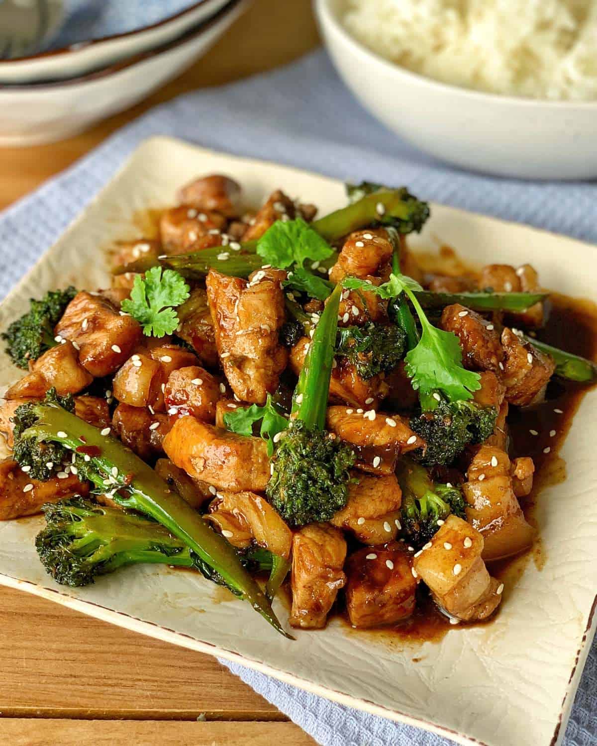 A plate of chicken and broccoli stir-fry garnished with cilantro and sesame seeds, served with a bowl of white rice in the background.