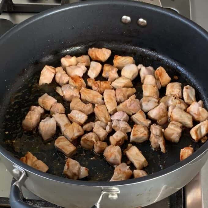 Cubed pieces of meat are browning in a large black skillet on a stovetop.