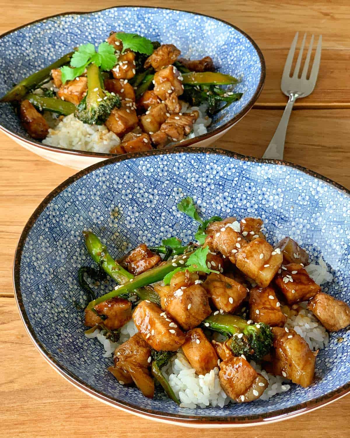 Two blue bowls filled with rice, stir-fried tofu, broccoli, green beans, and sesame seeds, garnished with cilantro, on a wooden table with a fork in the background.