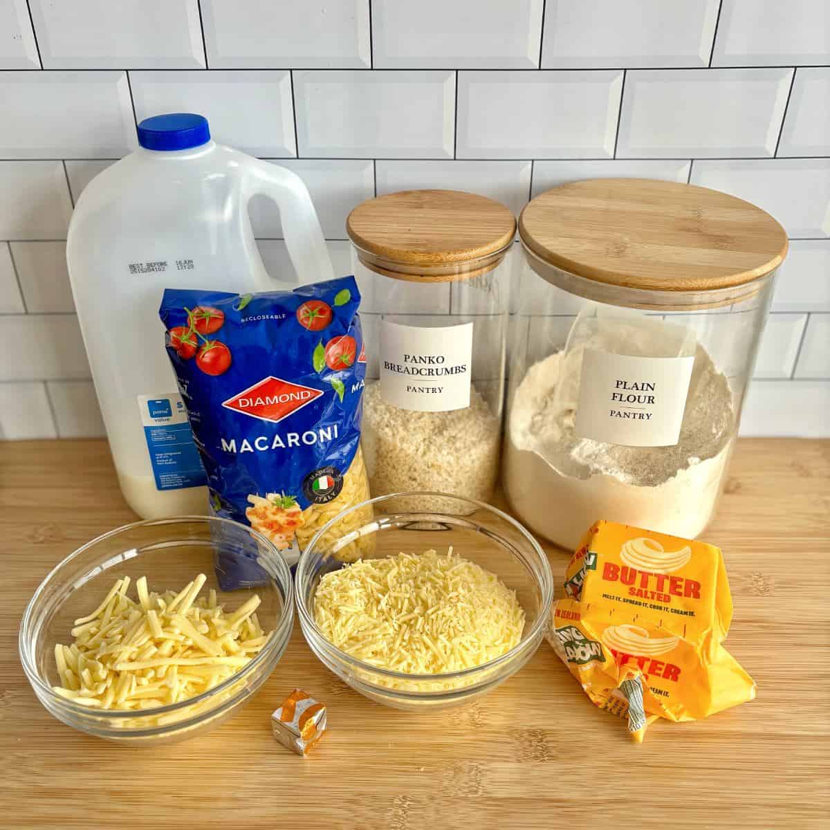 Slow Cooker Mac and Cheese Ingredients on a kitchen counter include milk, macaroni, panko breadcrumbs, plain flour, shredded cheese, grated cheese, butter, and a stock cube, all in containers or bowls.