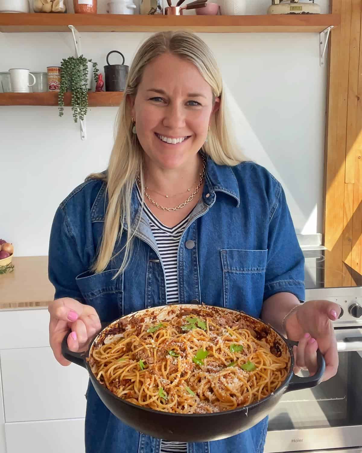 Lamb Ragu A woman in a denim shirt smiles while holding a pan of spaghetti topped with sauce and herbs in a modern kitchen.