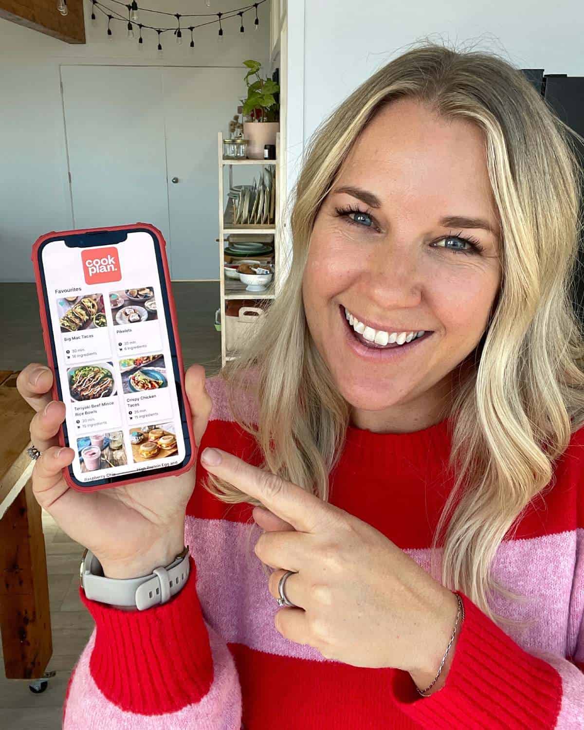 A woman in a red and pink sweater smiles and points at a smartphone displaying a recipe app called Cook Crate with various food images.