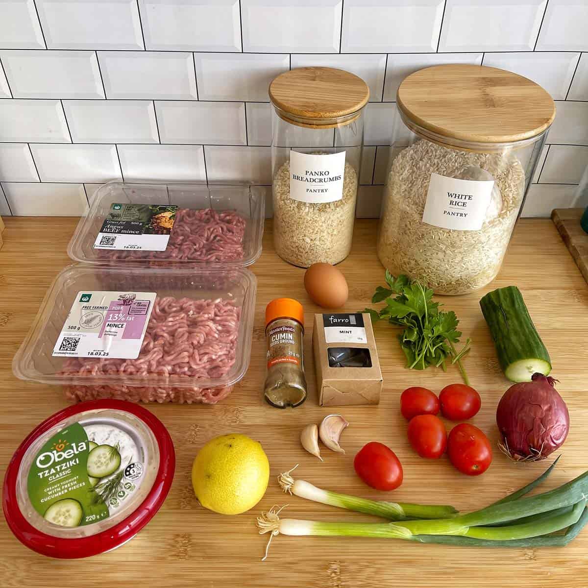 Assorted ingredients on a countertop including ground beef, white rice, panko breadcrumbs, an egg, herbs, vegetables, garlic, spices, lemon, and a container of tzatziki.