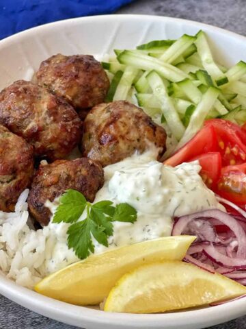A bowl with meatballs, white rice, sliced cucumber, tomato wedges, red onion, a dollop of tzatziki, a lemon wedge, and a parsley garnish.