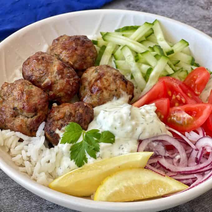 A bowl with meatballs, white rice, sliced cucumber, tomato wedges, red onion, a dollop of tzatziki, a lemon wedge, and a parsley garnish.