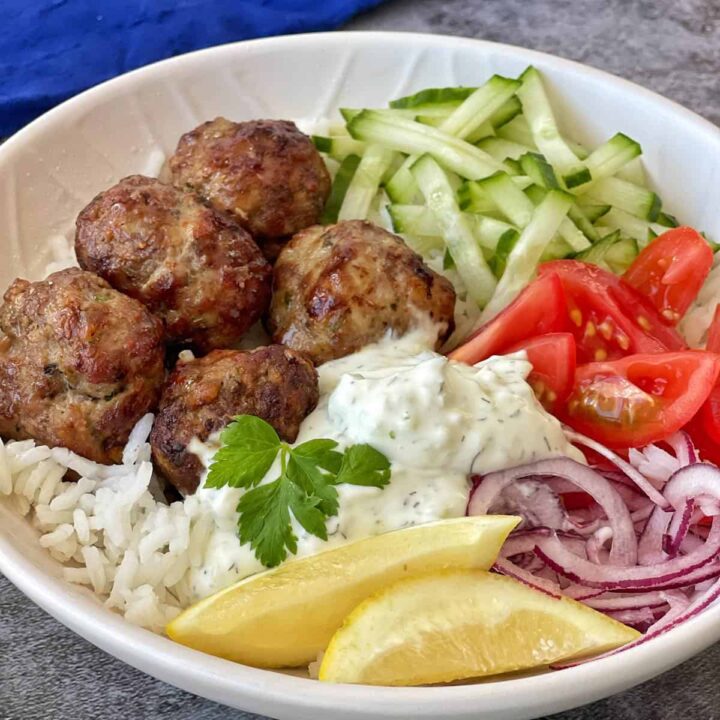 A bowl with meatballs, white rice, sliced cucumber, tomato wedges, red onion, a dollop of tzatziki, a lemon wedge, and a parsley garnish.