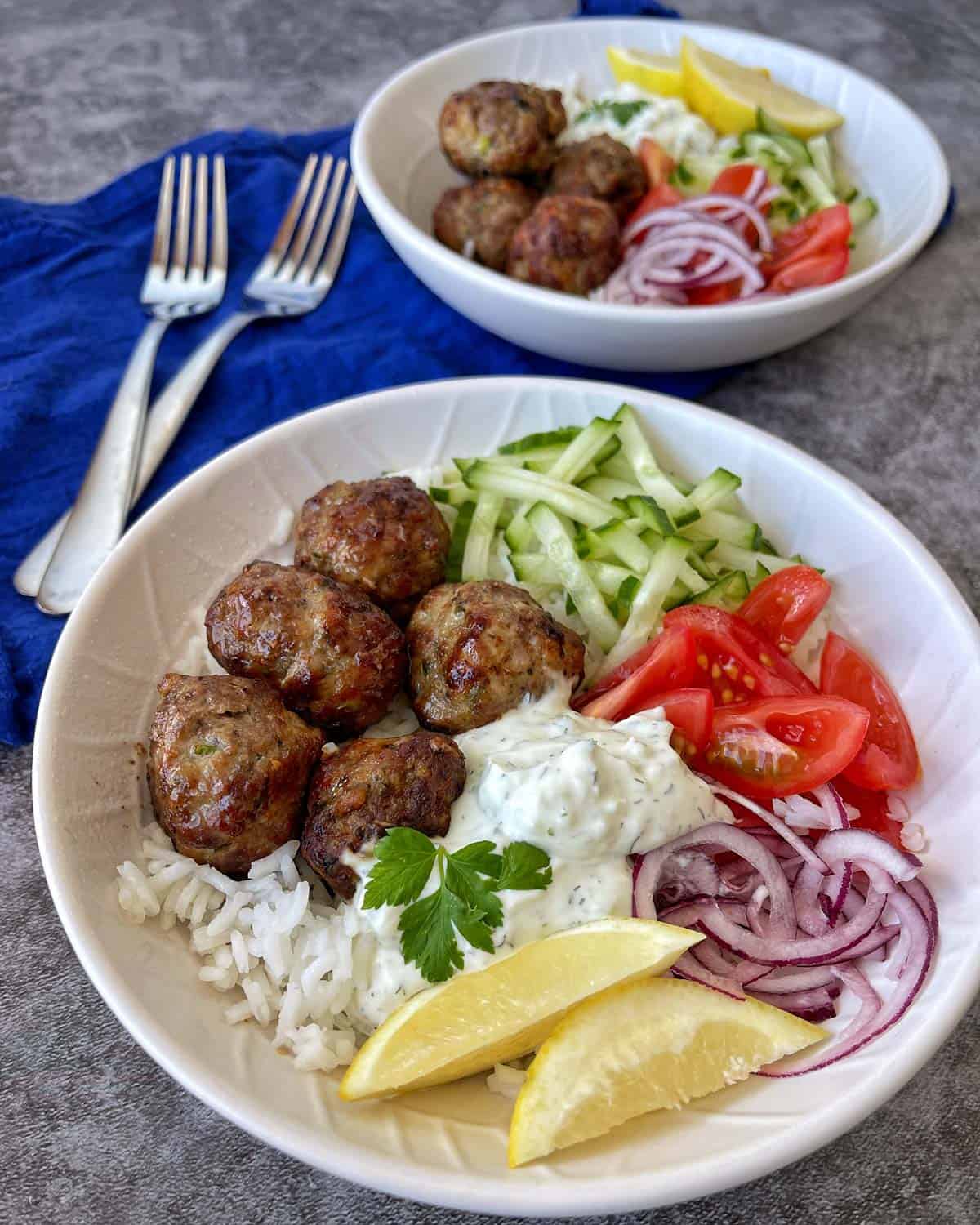 Two bowls of rice topped with meatballs, sliced cucumbers, tomatoes, red onions, lemon wedges, and a dollop of white sauce, placed on a gray surface with blue napkin and forks.