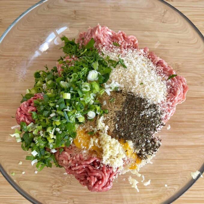 A glass bowl containing ground meat, chopped green onions, fresh herbs, panko breadcrumbs, grated garlic, ground spices, and a cracked egg, ready to be mixed.