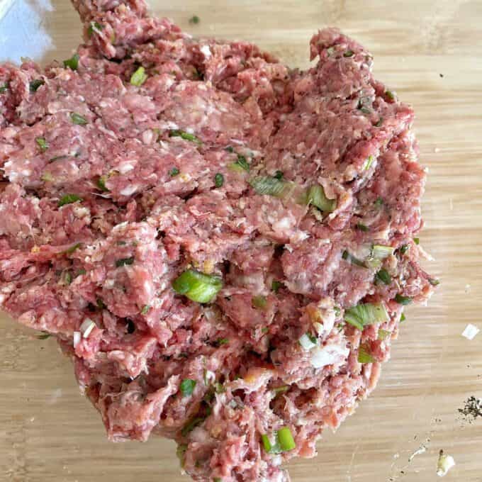 Raw ground meat mixed with chopped green onions and seasonings on a wooden surface.