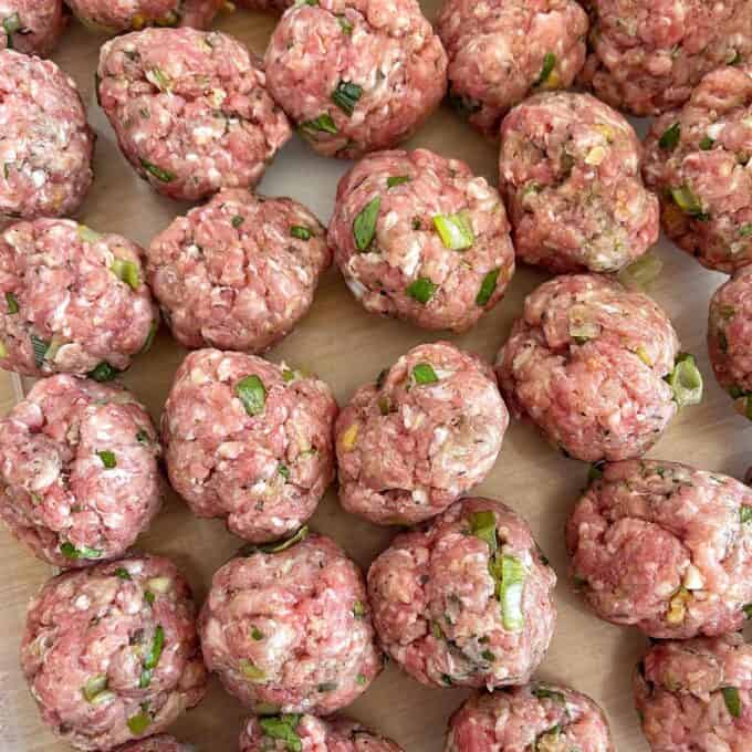 Uncooked meatballs mixed with chopped green onions and herbs arranged on a flat surface.
