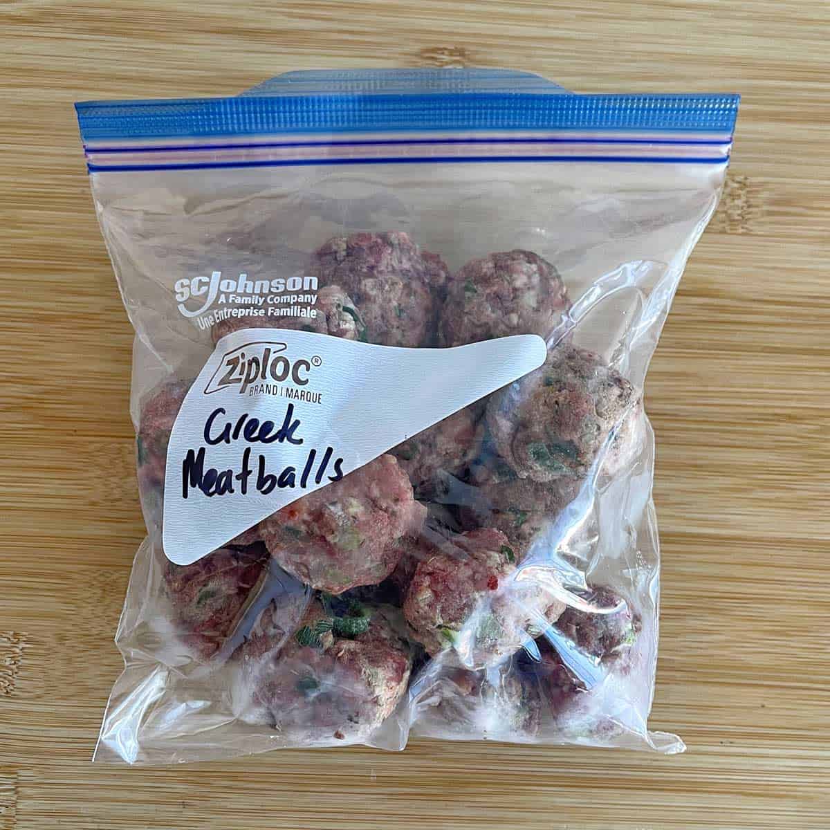 A clear Ziploc bag labeled Greek Meatballs filled with uncooked meatballs sits on a wooden surface.