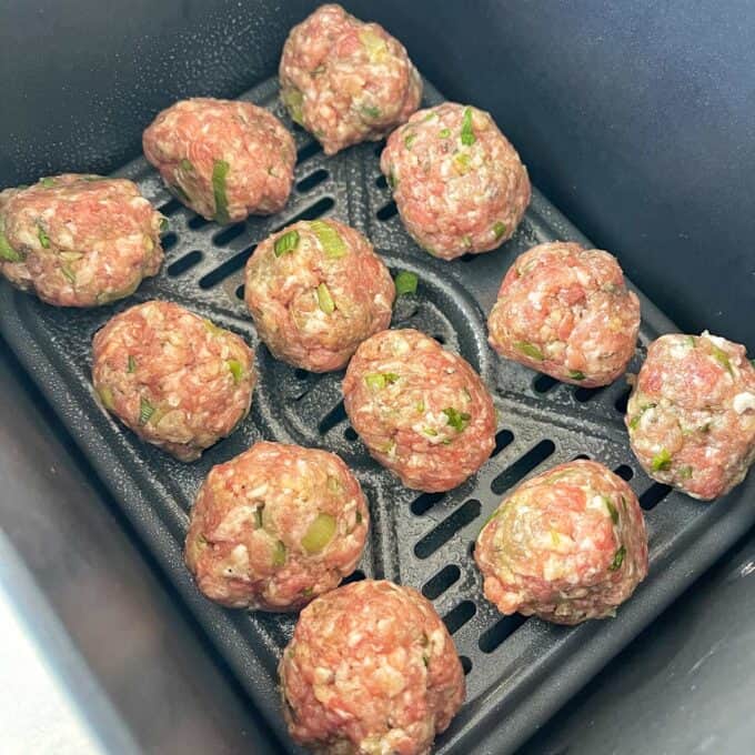 Raw meatballs with chopped green herbs are arranged in a single layer inside an air fryer basket, ready to be cooked.
