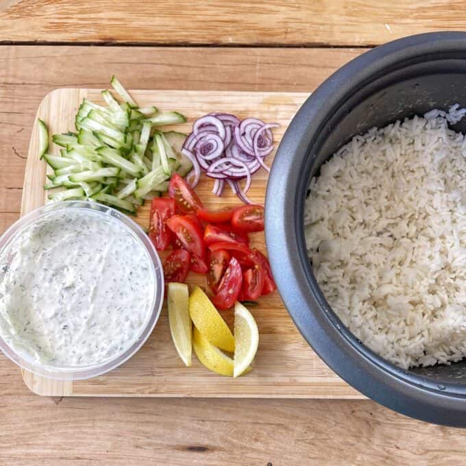 A rice cooker with white rice next to a cutting board holding sliced cucumber, red onion, tomatoes, lemon wedges, and a container of creamy sauce.