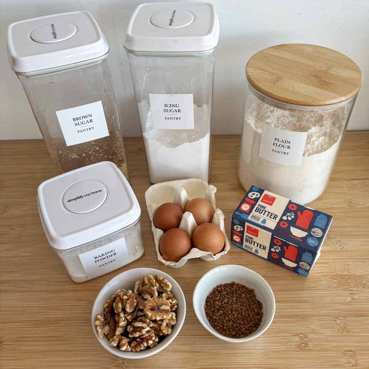 Containers of brown sugar, icing sugar, plain flour, and walnuts, a carton of eggs, a block of butter, and a bowl of coffee granules arranged on a wooden surface.