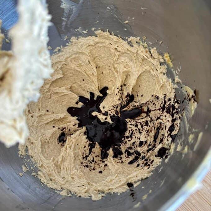 A mixing bowl with creamed butter and sugar, partially mixed with a dark liquid, likely vanilla extract, in the center.