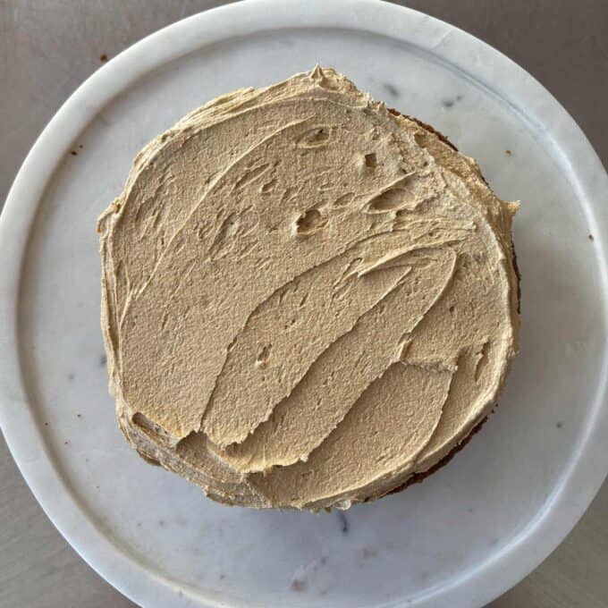 A round cake with light brown frosting is displayed on a white marble platter, viewed from above.
