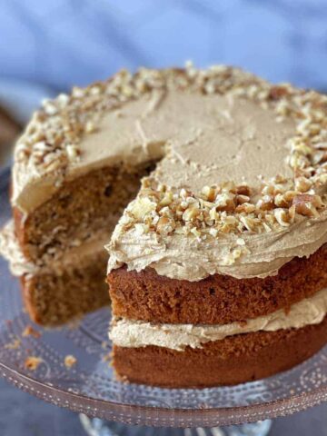 A two-layer cake with light brown frosting and chopped nuts on top, displayed on a glass cake stand with a slice removed.