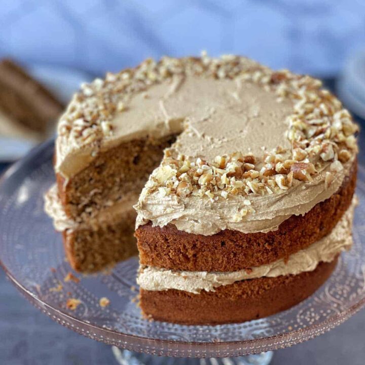 A two-layer cake with light brown frosting and chopped nuts on top, displayed on a glass cake stand with a slice removed.