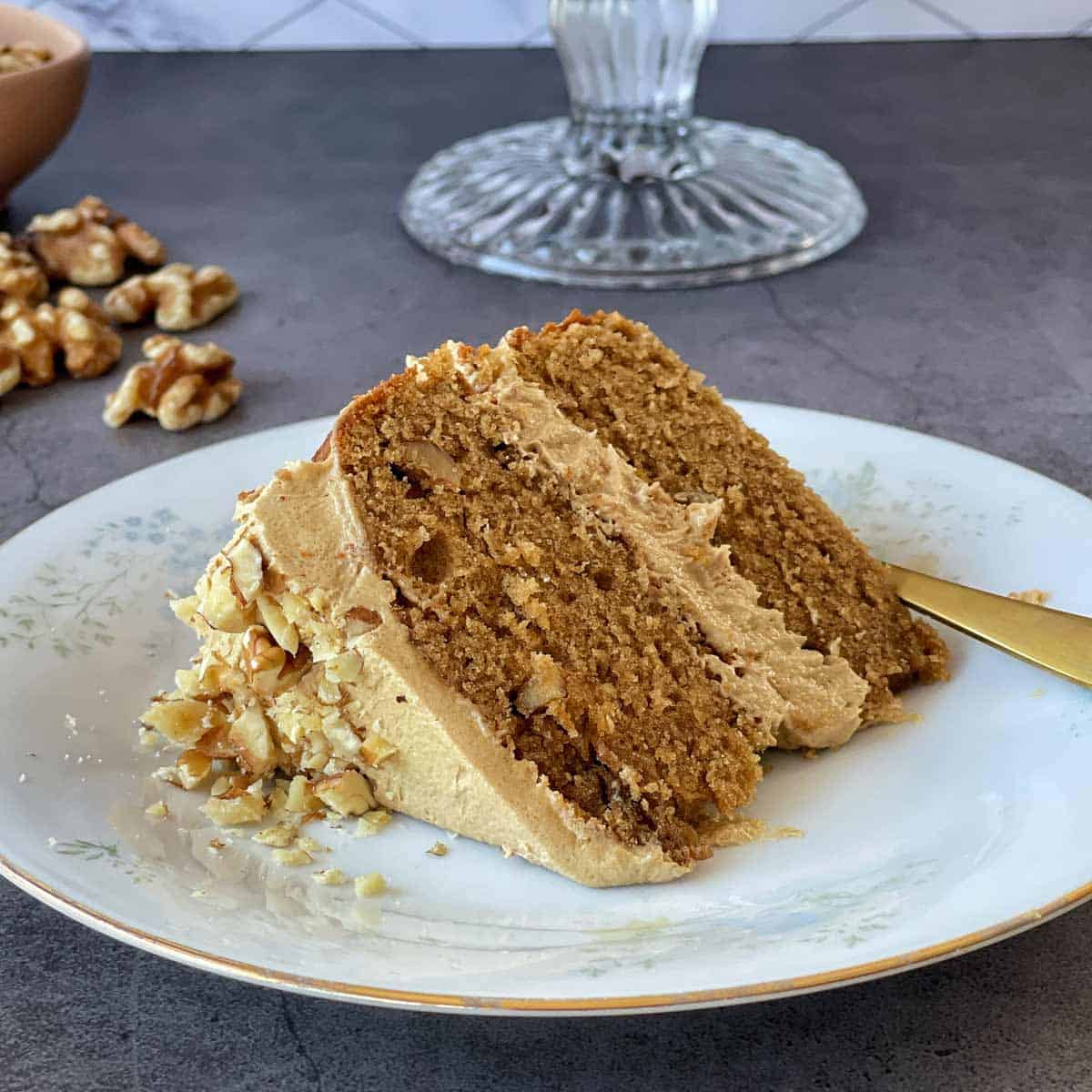 A slice of walnut cake with light brown frosting and chopped walnuts on a white plate, with a gold fork beside it. Walnuts and an empty cake stand are in the background.