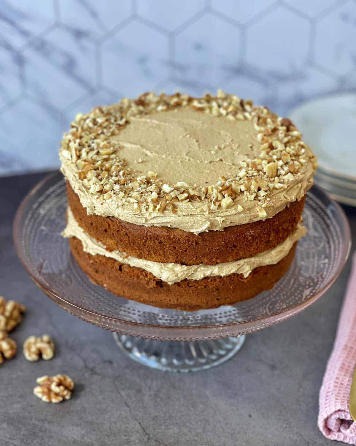 A two-layer cake with light brown frosting and chopped walnuts on top, displayed on a glass cake stand against a tiled background.